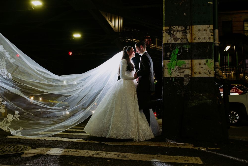 A bride and groom share an intimate moment under dramatic lighting while her long veil flows in the evening breeze.