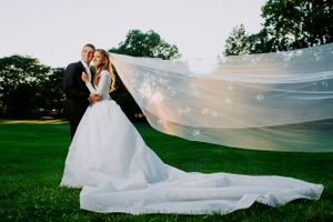Wedding photo in green field with dramatic flowing veil catching the wind.