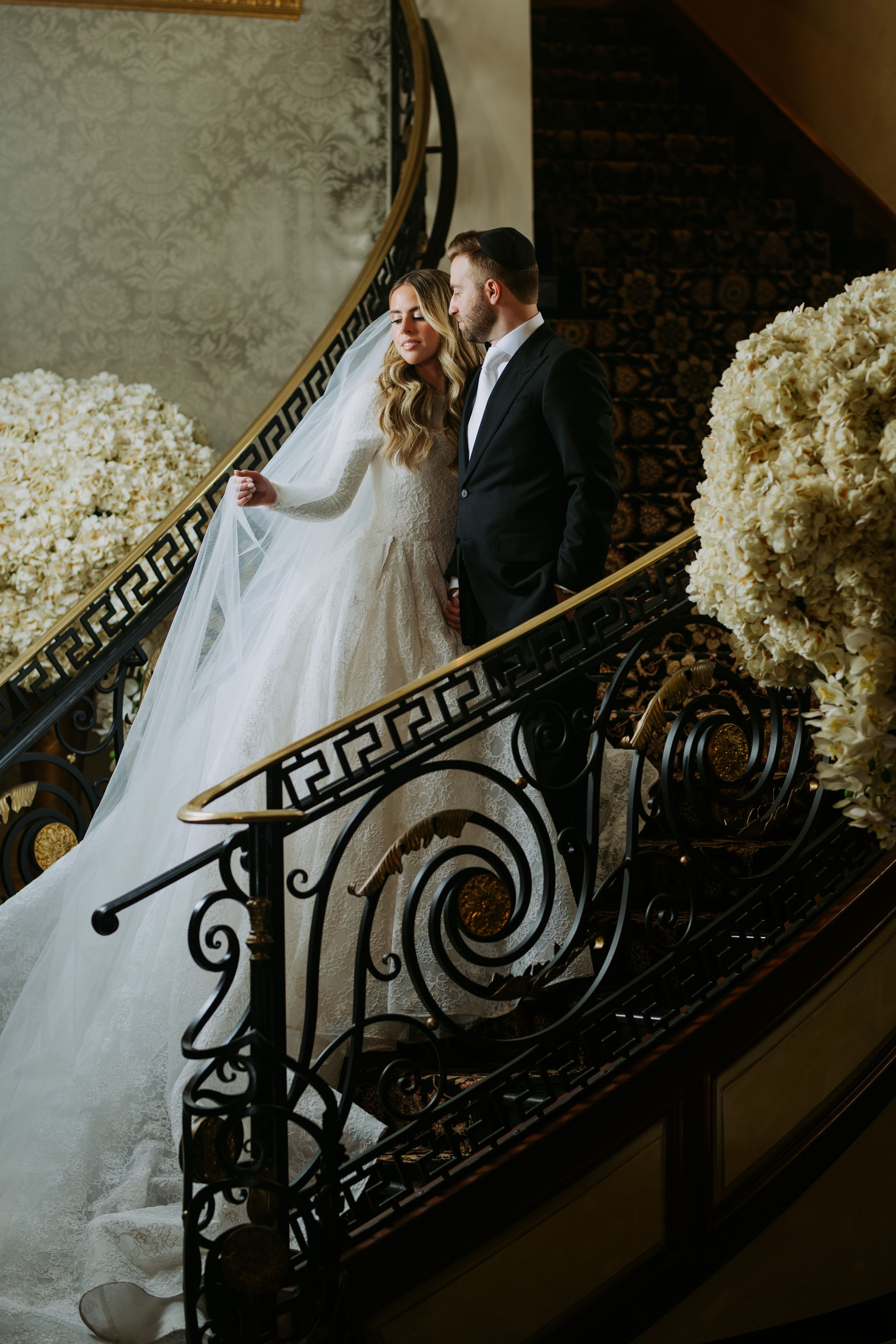 Elegant couple on ornate staircase with decorative iron railings and white floral arrangements.