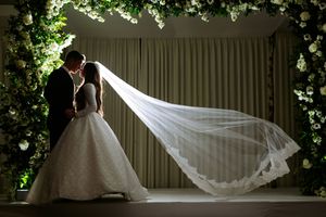 A flowing white wedding veil billows dramatically in the breeze against a natural outdoor backdrop with greenery.