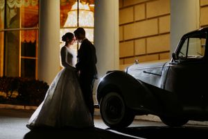 Silhouetted wedding couple posing beside vintage car in evening light.