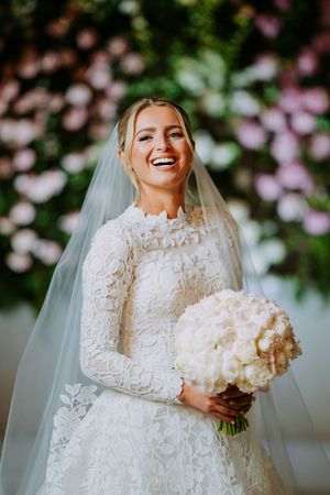 A joyful bride in a long-sleeved lace wedding gown holds a white bouquet while laughing.
