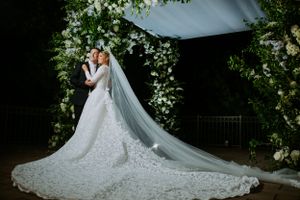 Romantic wedding scene under a floral arch with a dramatic flowing cathedral veil and lace gown.