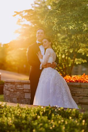 A couple in wedding attire embraces during golden hour sunset near a stone wall and vibrant orange flowers.