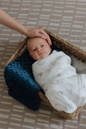 Baby swaddled in white blanket rests in woven basket with blue pillow.