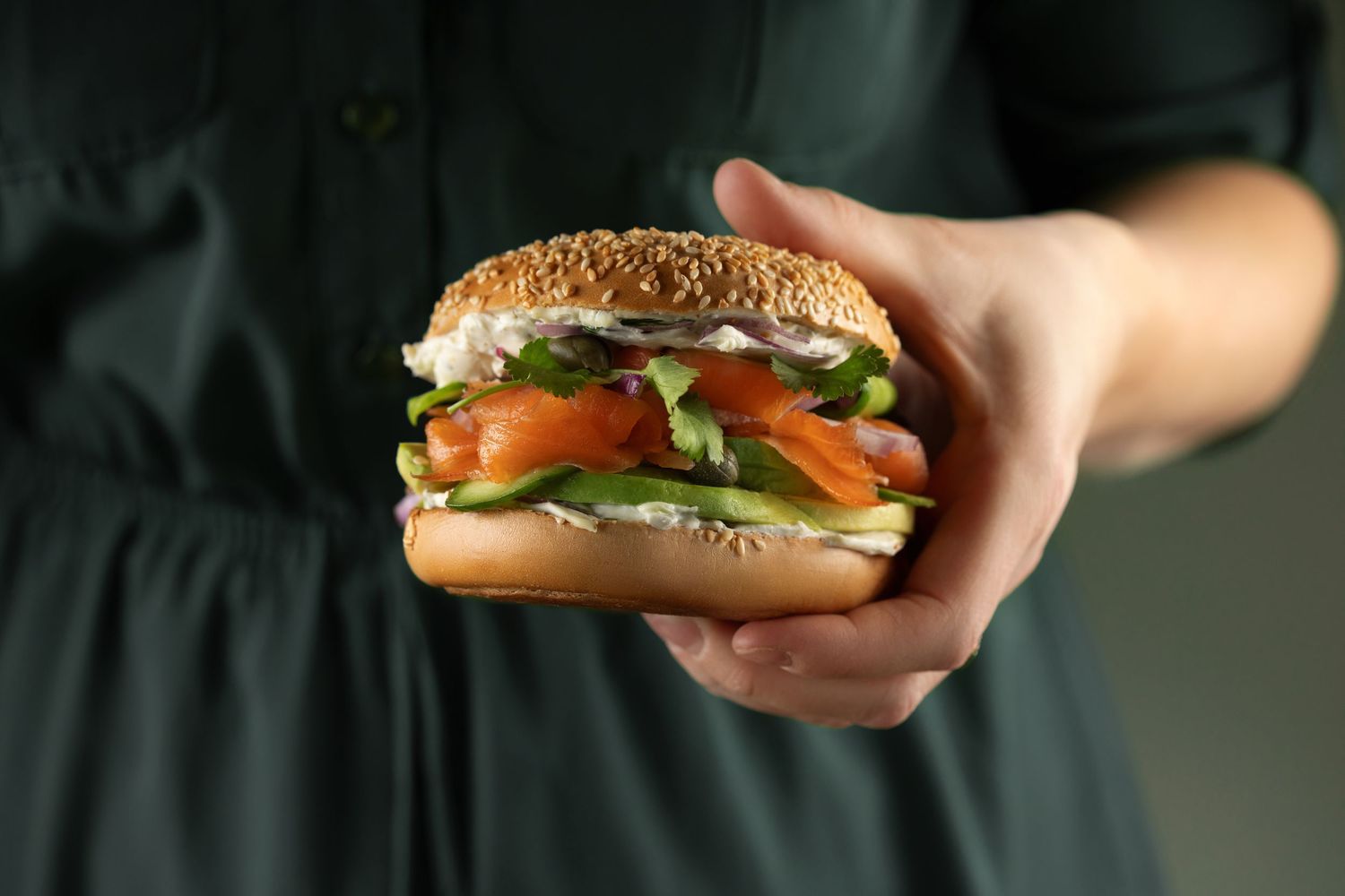 Hands holding a fresh sandwich with lettuce, tomatoes and cheese against a dark background.