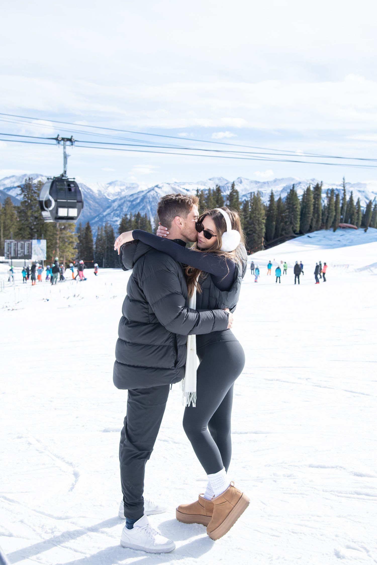 Chic young couple stands together with a ski gondola in the background on top of Aspen ski mountain during an engagement photo session.