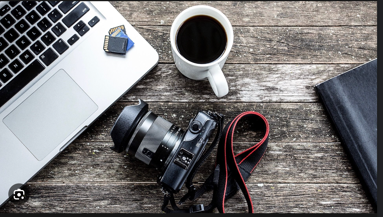 Modern workspace with laptop, coffee cup, and camera equipment on weathered wood surface.
