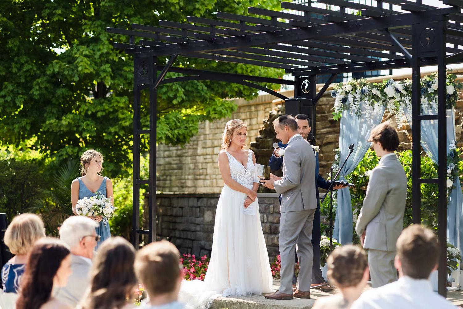 An outdoor wedding ceremony takes place under a black pergola surrounded by lush green trees and natural light in Columbus Ohio