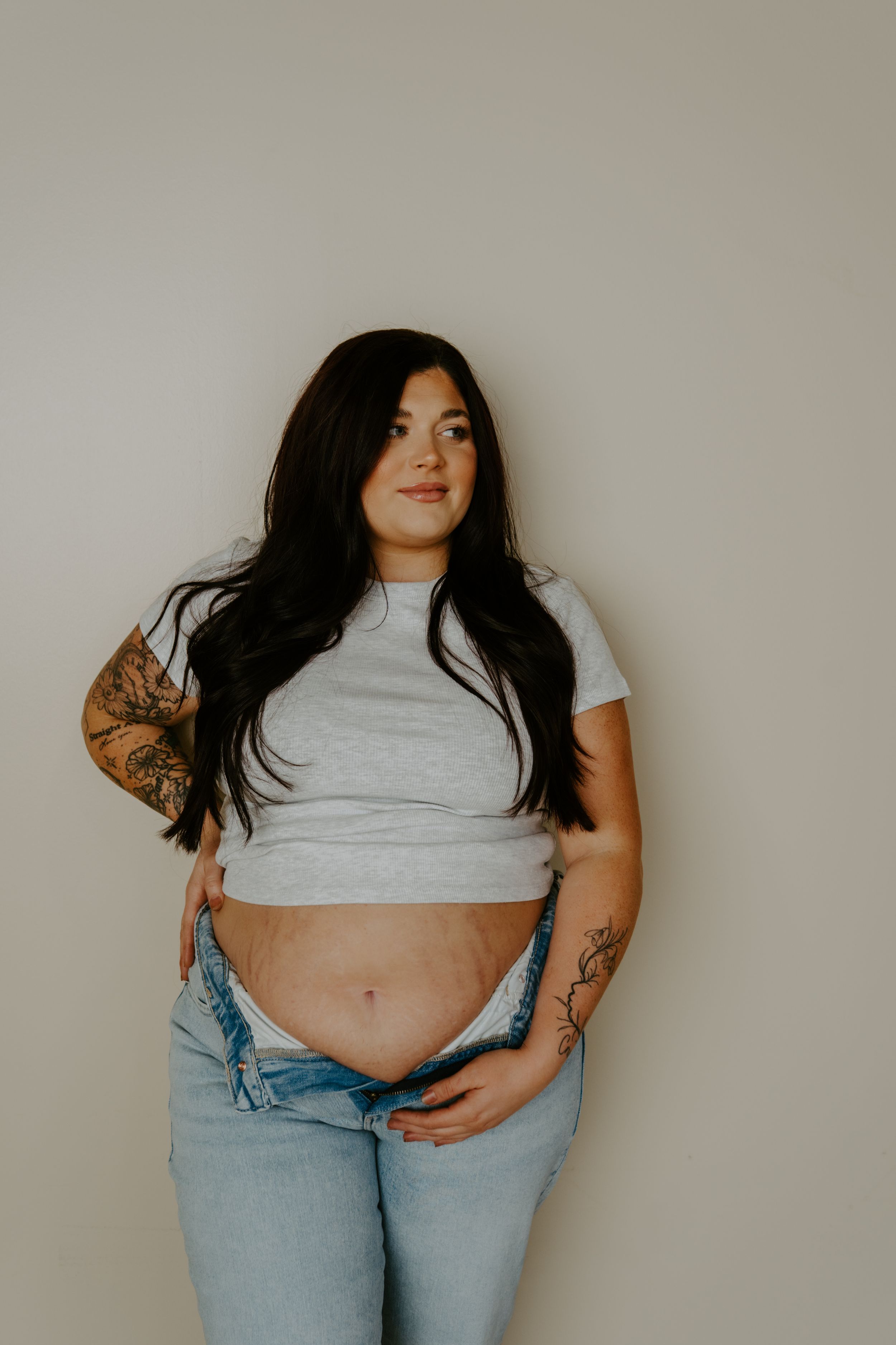 A woman in casual attire poses against a neutral wall showing her midriff and tattoos.