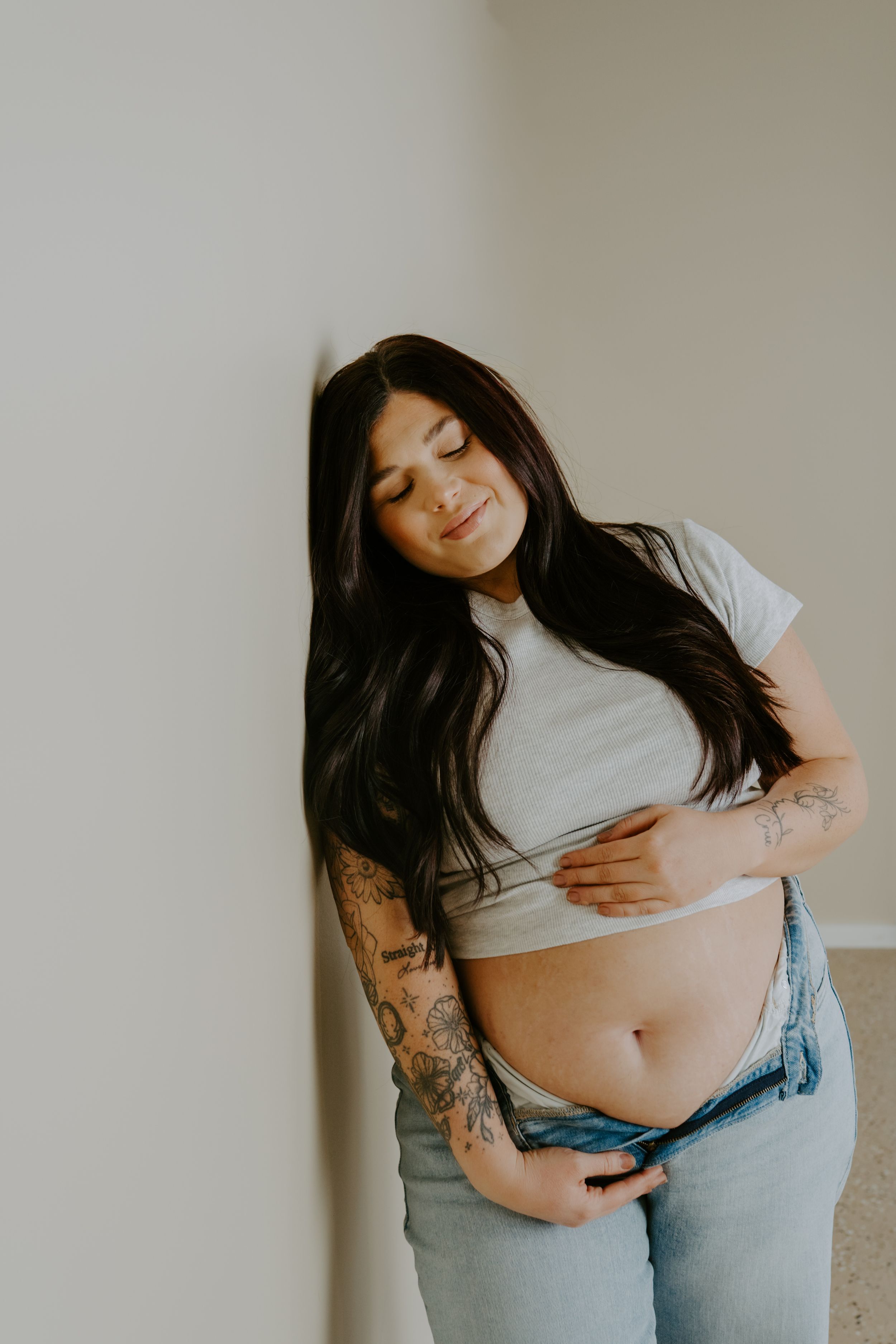 A woman in denim and crop top poses against a neutral wall showing her midriff.