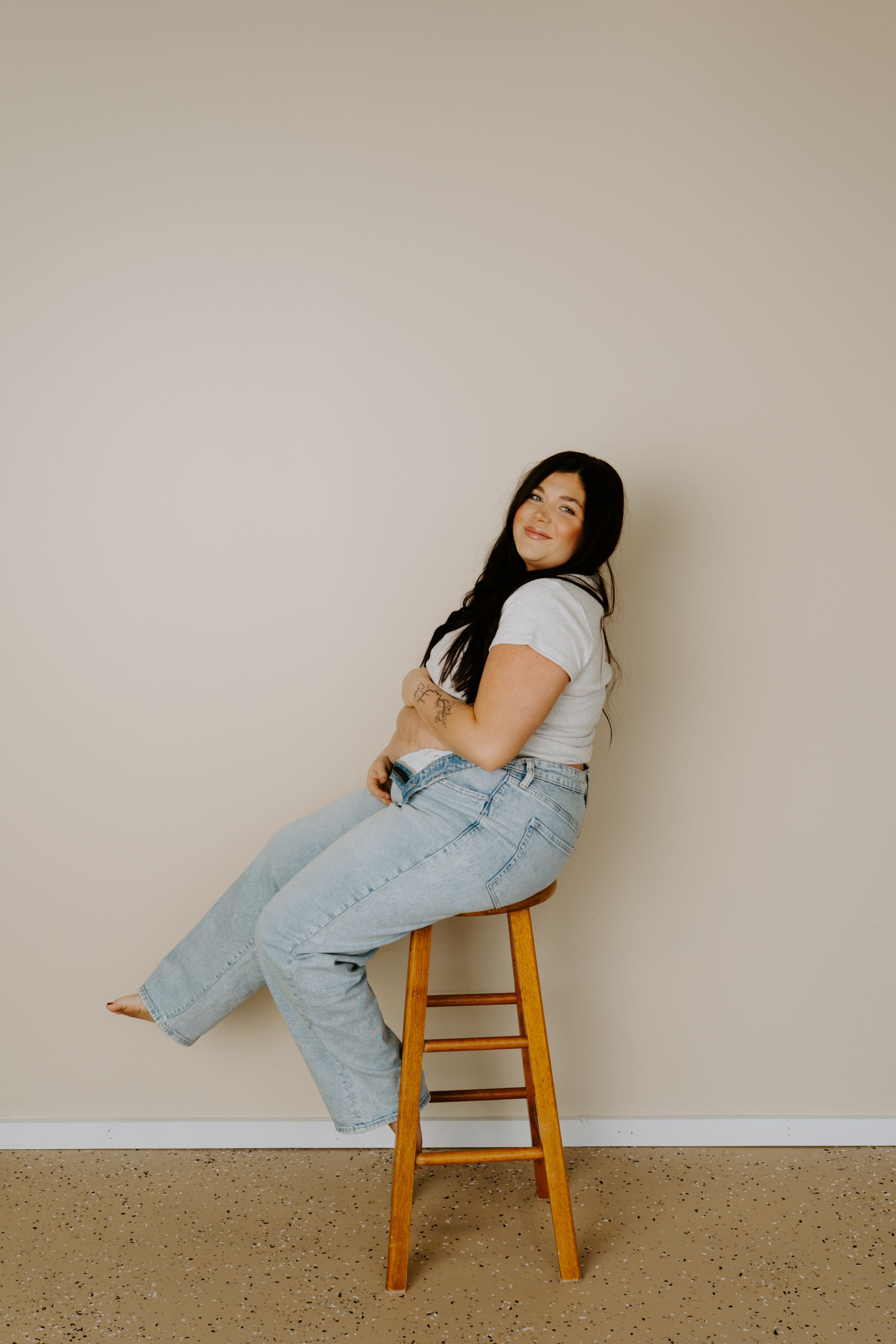 A woman in casual attire poses on a wooden stool against a neutral wall.
