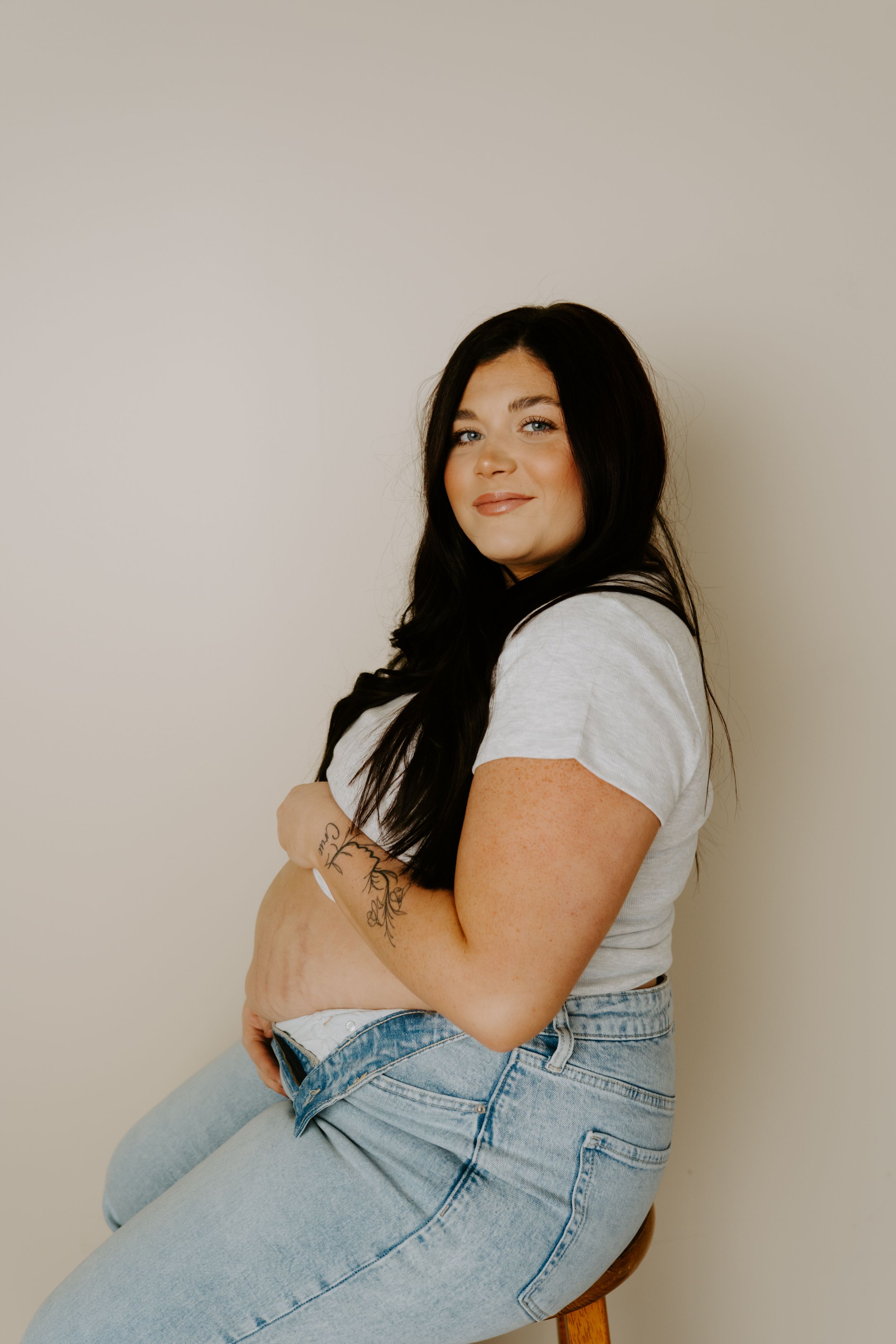 A woman in light denim and cropped shirt poses on a wooden stool against a neutral wall.