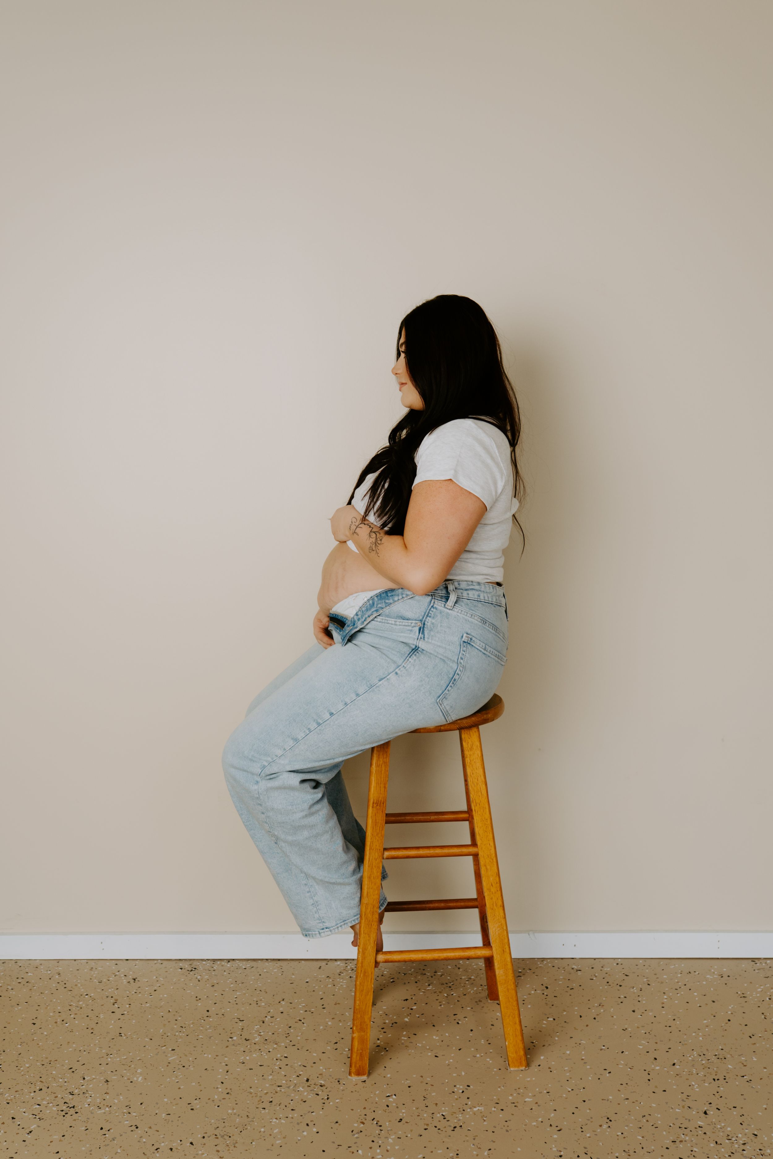 A side profile of a woman sitting on a wooden stool against a neutral wall.