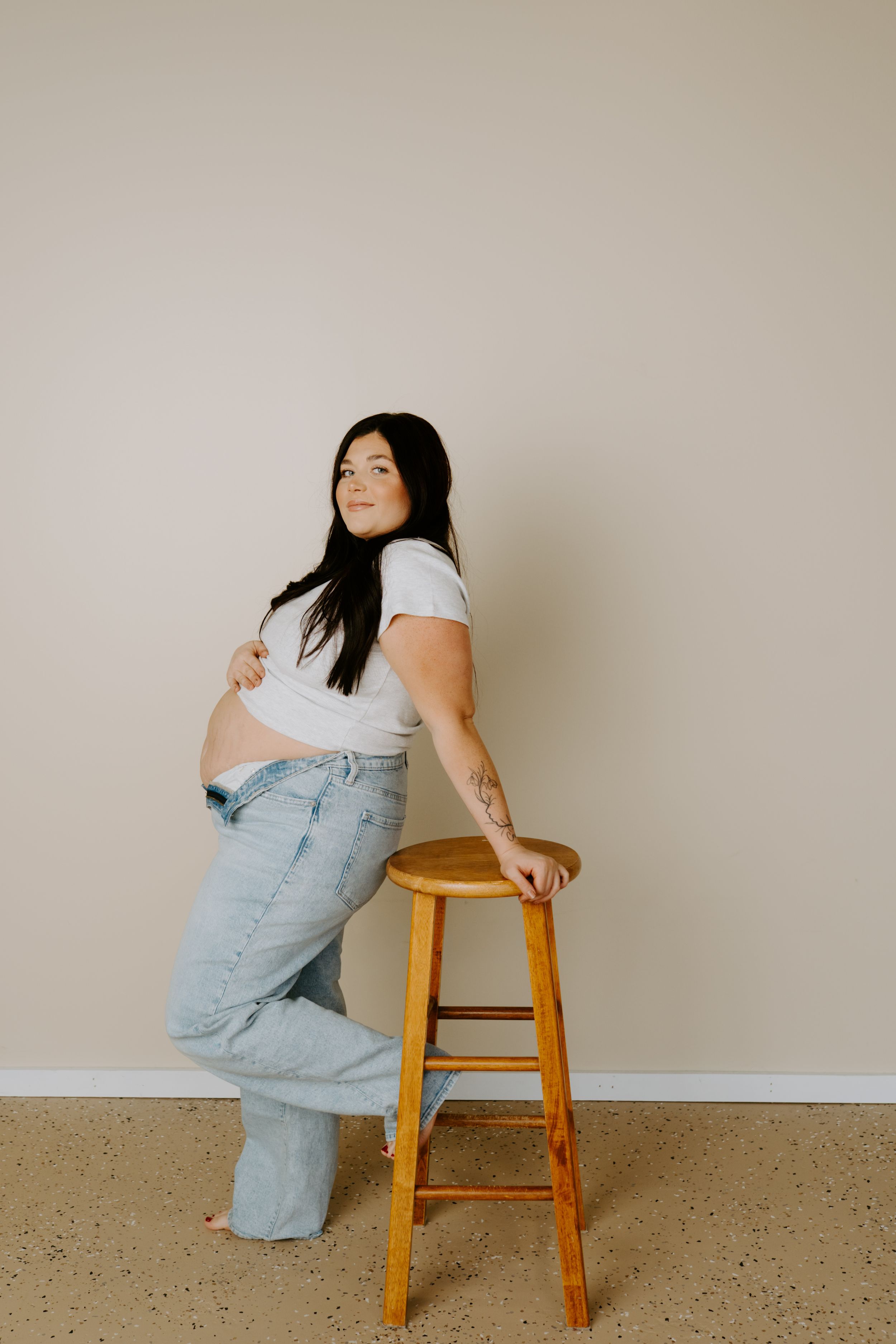 Person in light denim overalls and white t-shirt poses casually with wooden stool against neutral wall.