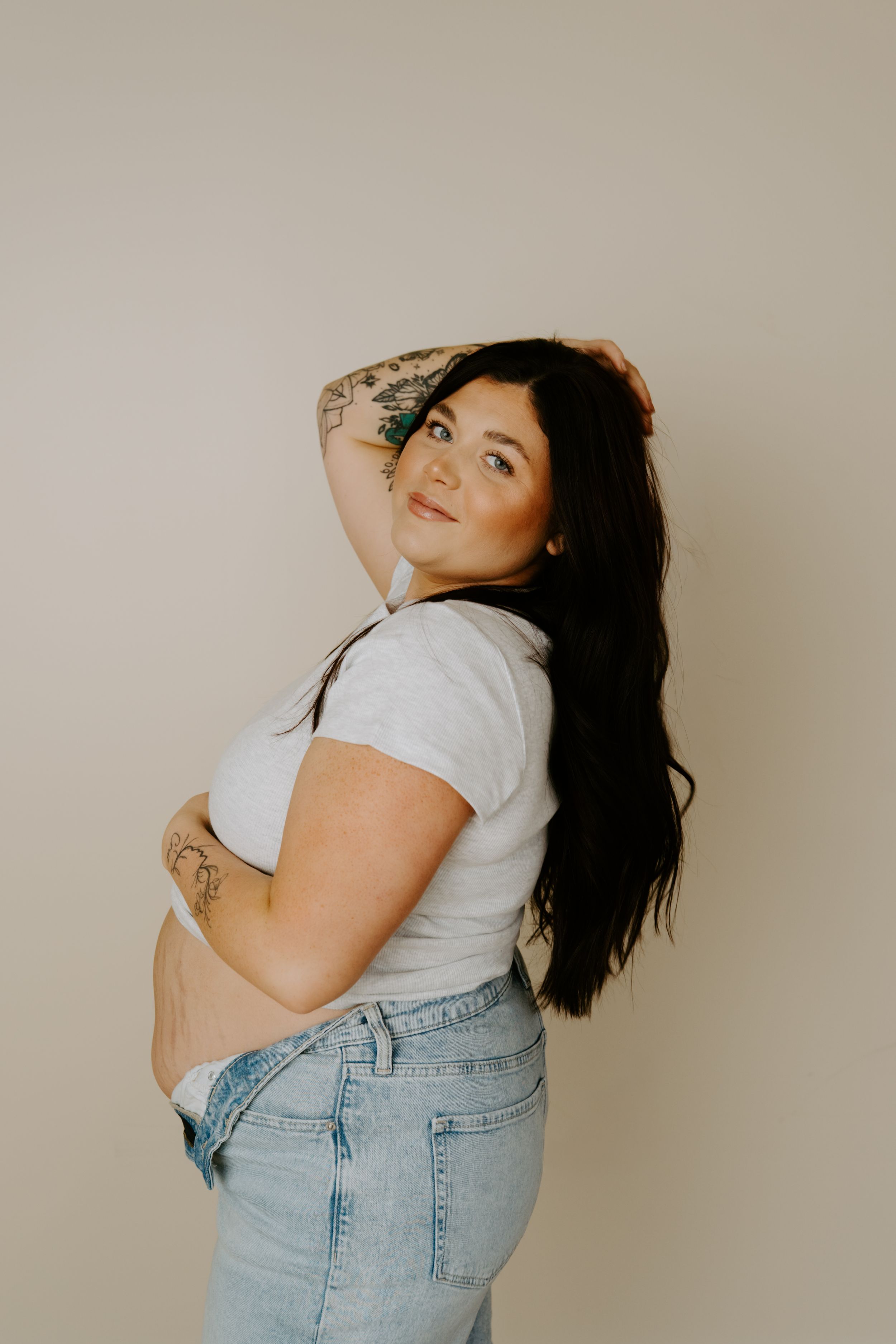 Model wearing denim overalls and white shirt poses expressively against beige backdrop.