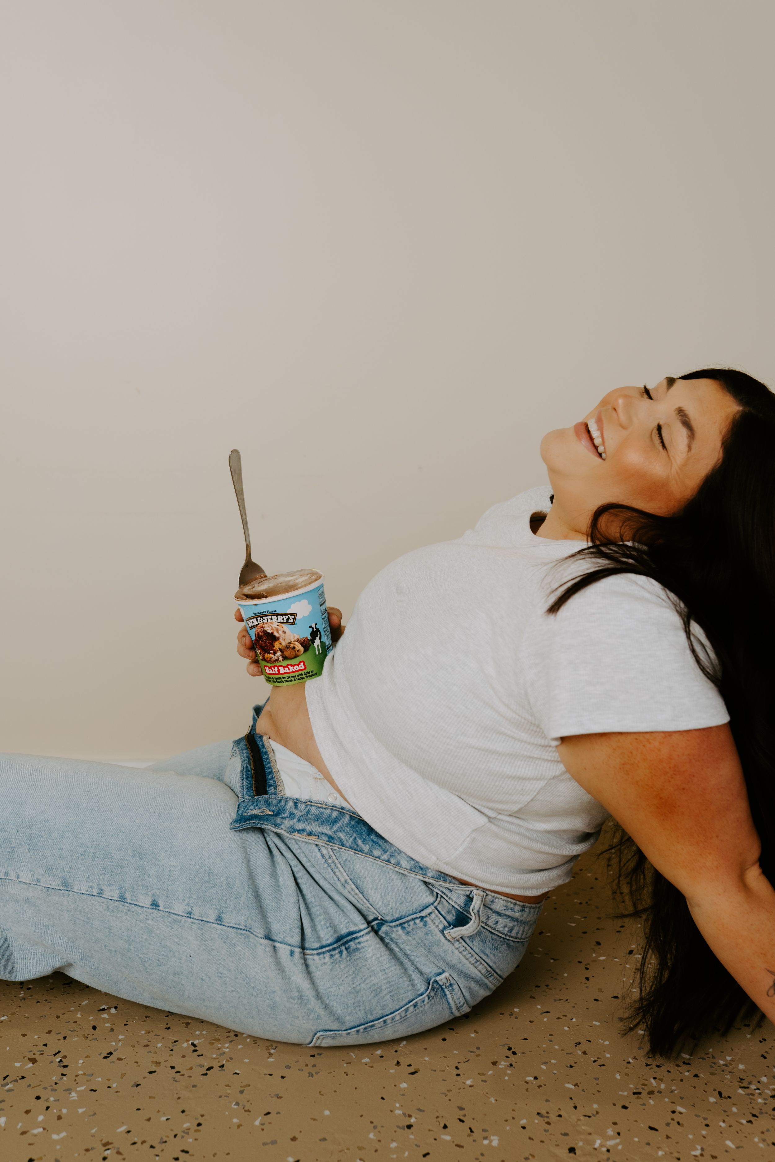 Individual lying on floor with ice cream container in relaxed pose wearing denim and white top.