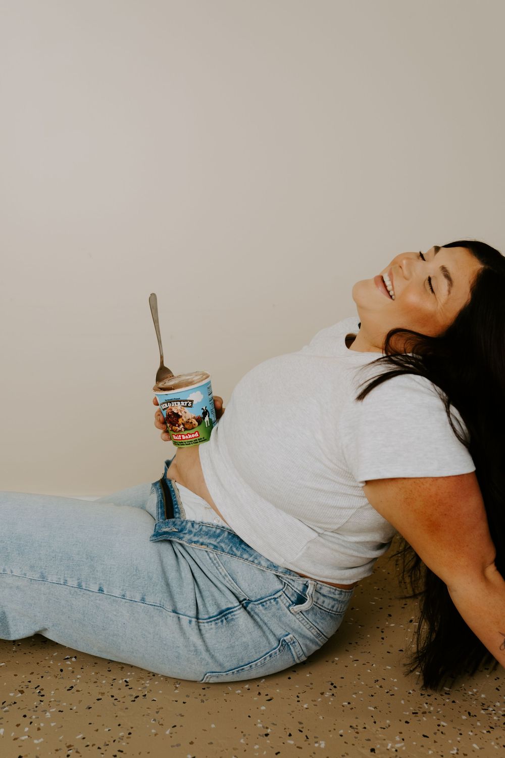Individual lying on floor with ice cream container in relaxed pose wearing denim and white top.