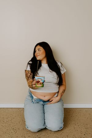 Person sits cross-legged on floor with ice cream container wearing casual denim and white outfit.