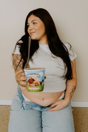 Figure in denim overalls and white top poses with ice cream container against neutral background.