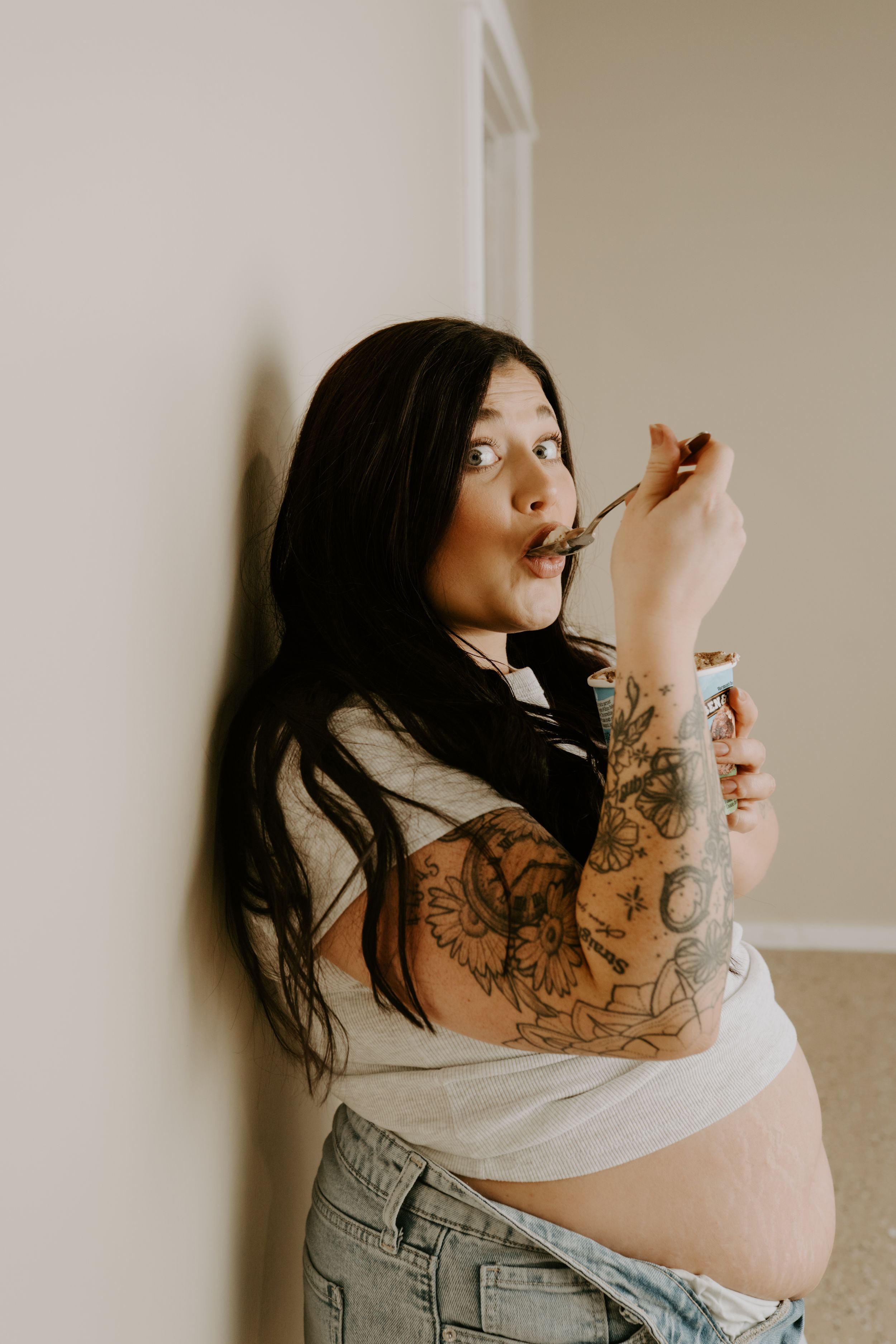 Individual with tattoos poses with ice cream container wearing denim overalls and cropped top.