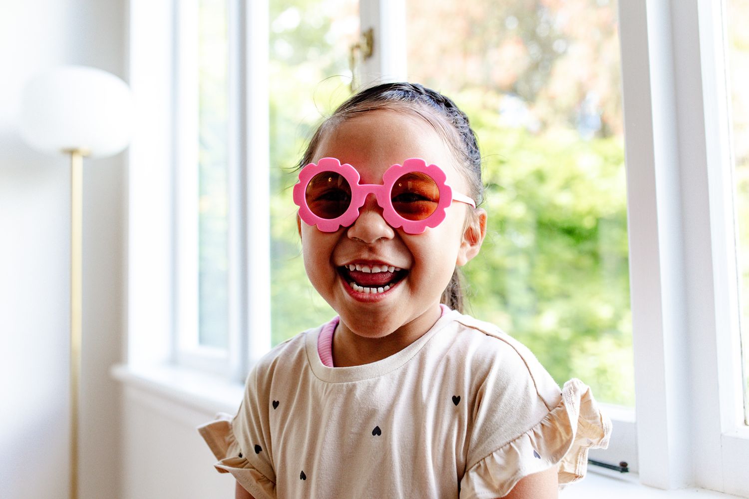 Two people wearing pink heart-shaped sunglasses laugh together in a bright sunlit room.