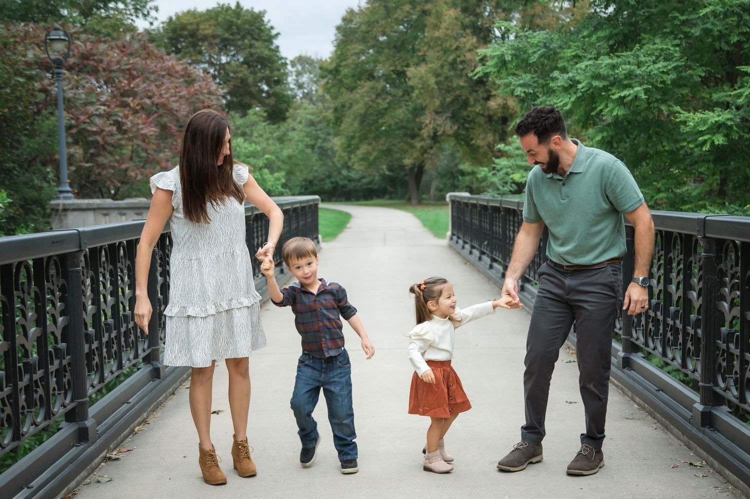 A family walks together holding hands on a scenic bridge path surrounded by lush green trees.