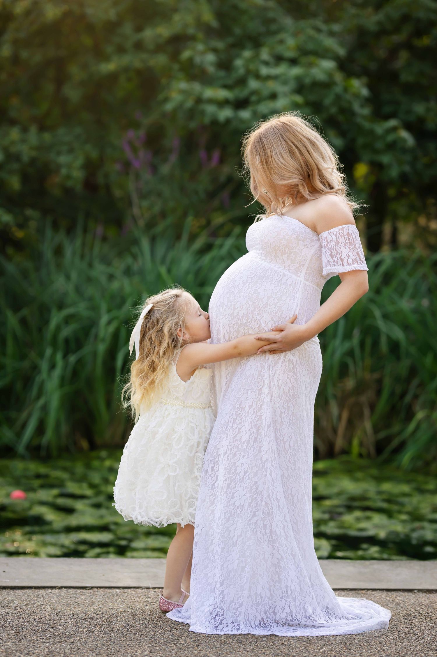 Mother and daughter in matching white lace dresses share tender moments by a garden pond.