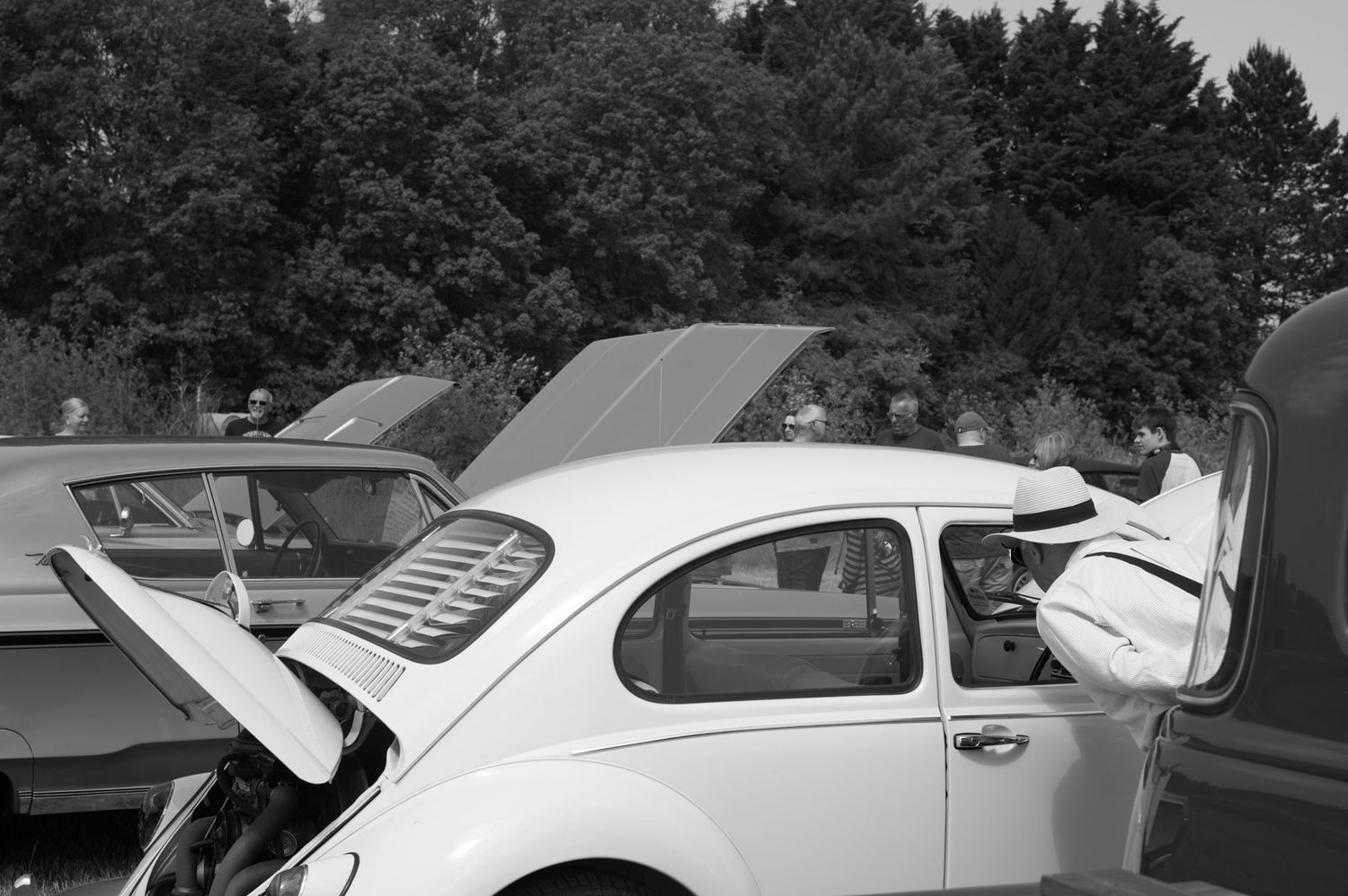 A classic Volkswagen Beetle with its engine compartment open being inspected at a car show or gathering.
