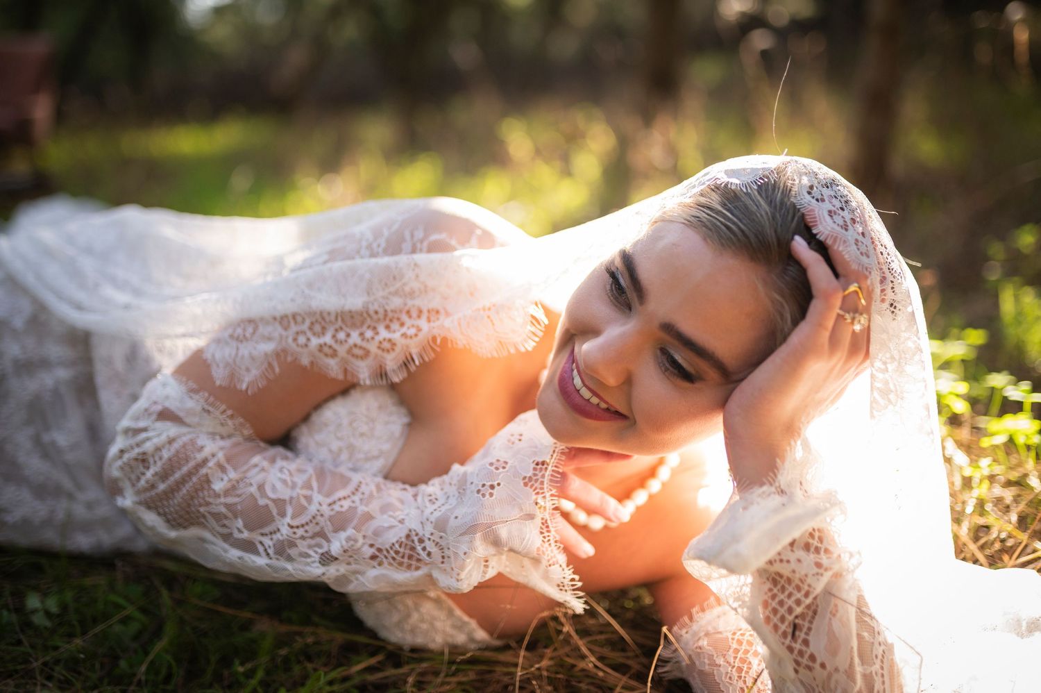 A bride in a white lace wedding dress lies gracefully in golden sunlit grass during an outdoor photo session.