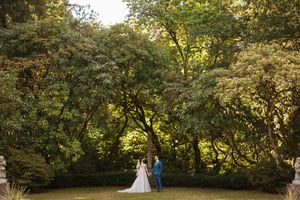 A wedding couple walks through a lush garden pathway surrounded by green trees during golden hour.