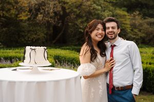 A couple stands together outdoors next to their elegant white wedding cake on a table during a garden celebration.