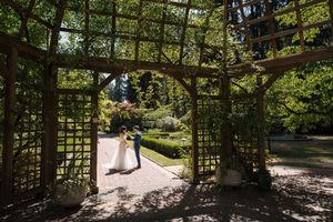 A couple in wedding attire stands under a wooden garden arbor surrounded by lush greenery on a sunny day.