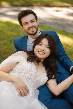 A couple in wedding attire relaxes on grass during an outdoor photo session, with soft natural lighting.