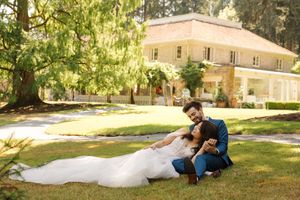 Newlyweds share intimate moments on a sunny lawn with historic building in background.