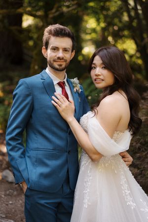 A couple in wedding attire poses together in a garden setting with natural sunlight filtering through the trees.