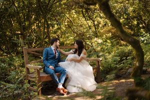 A couple in wedding attire sits together on a rustic wooden bridge surrounded by lush green forest.