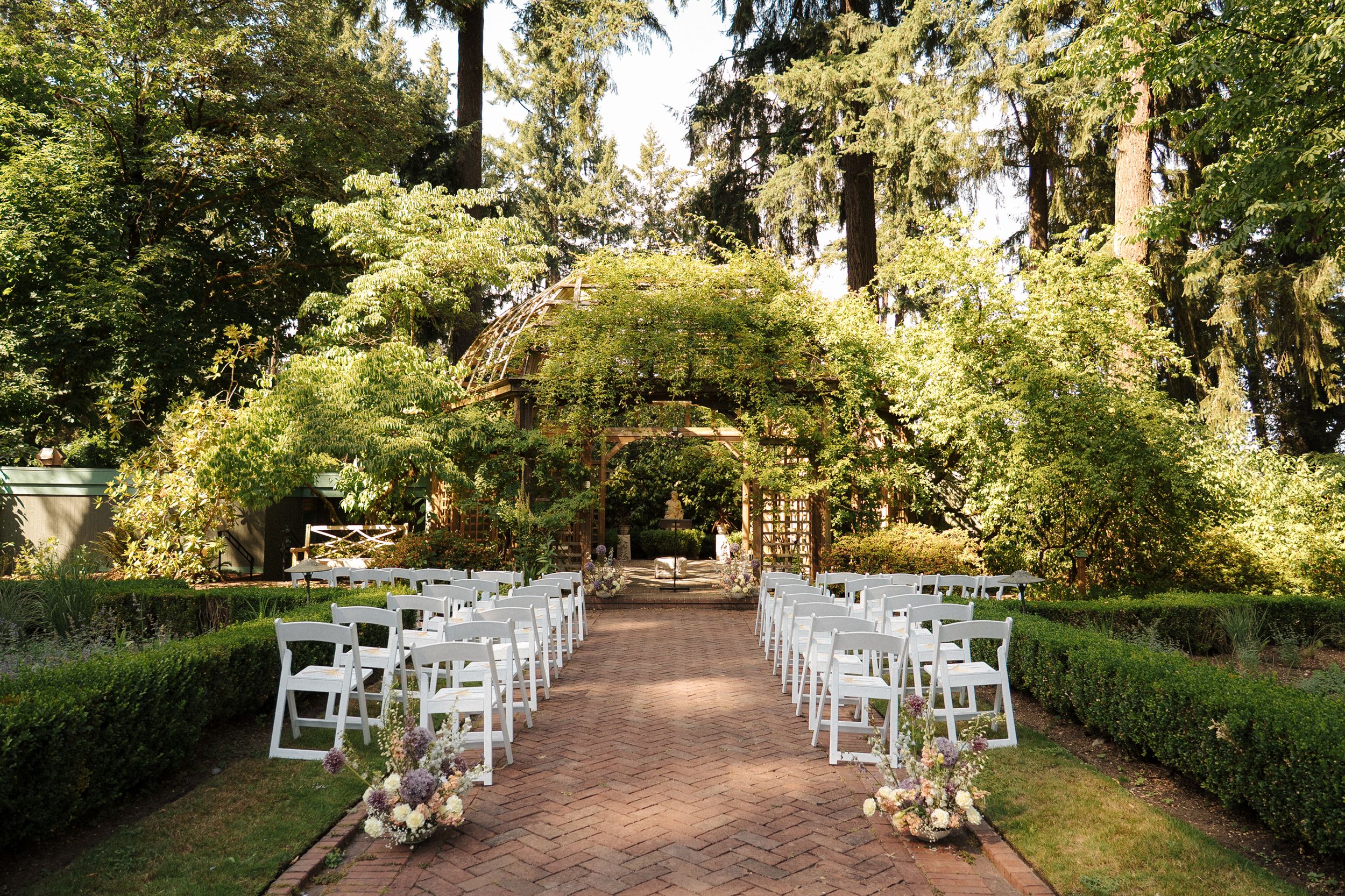 Garden wedding ceremony setup with white chairs lining a tree-covered pathway.