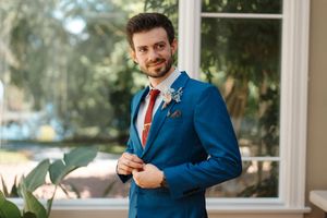 A person in a navy blue suit adjusts their tie while standing in front of a window.