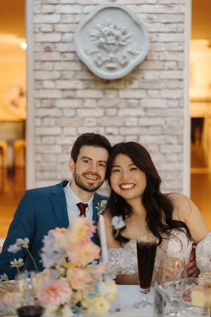A couple sits at an elegant wedding reception table with brick walls and floral centerpieces.