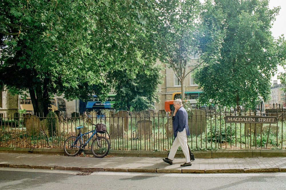 Person walking past iron fence and brick buildings with bicycle parked against railing.