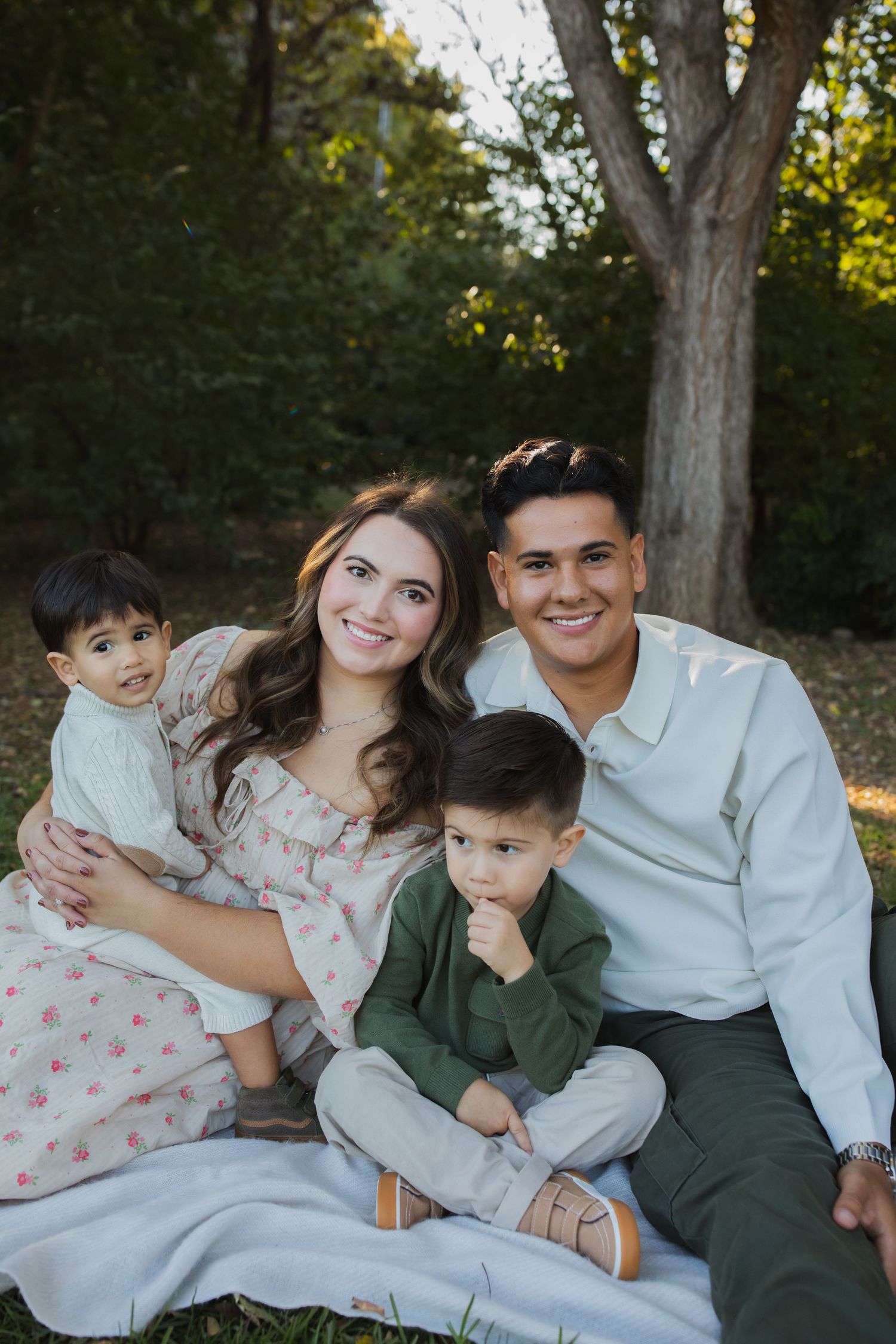 A family of four sits together on a blanket for an outdoor portrait session in a wooded setting.