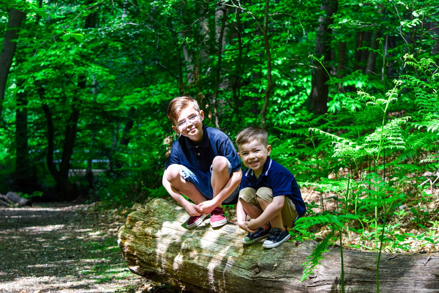 Two young boys sit together on a fallen log in a lush green forest setting.
