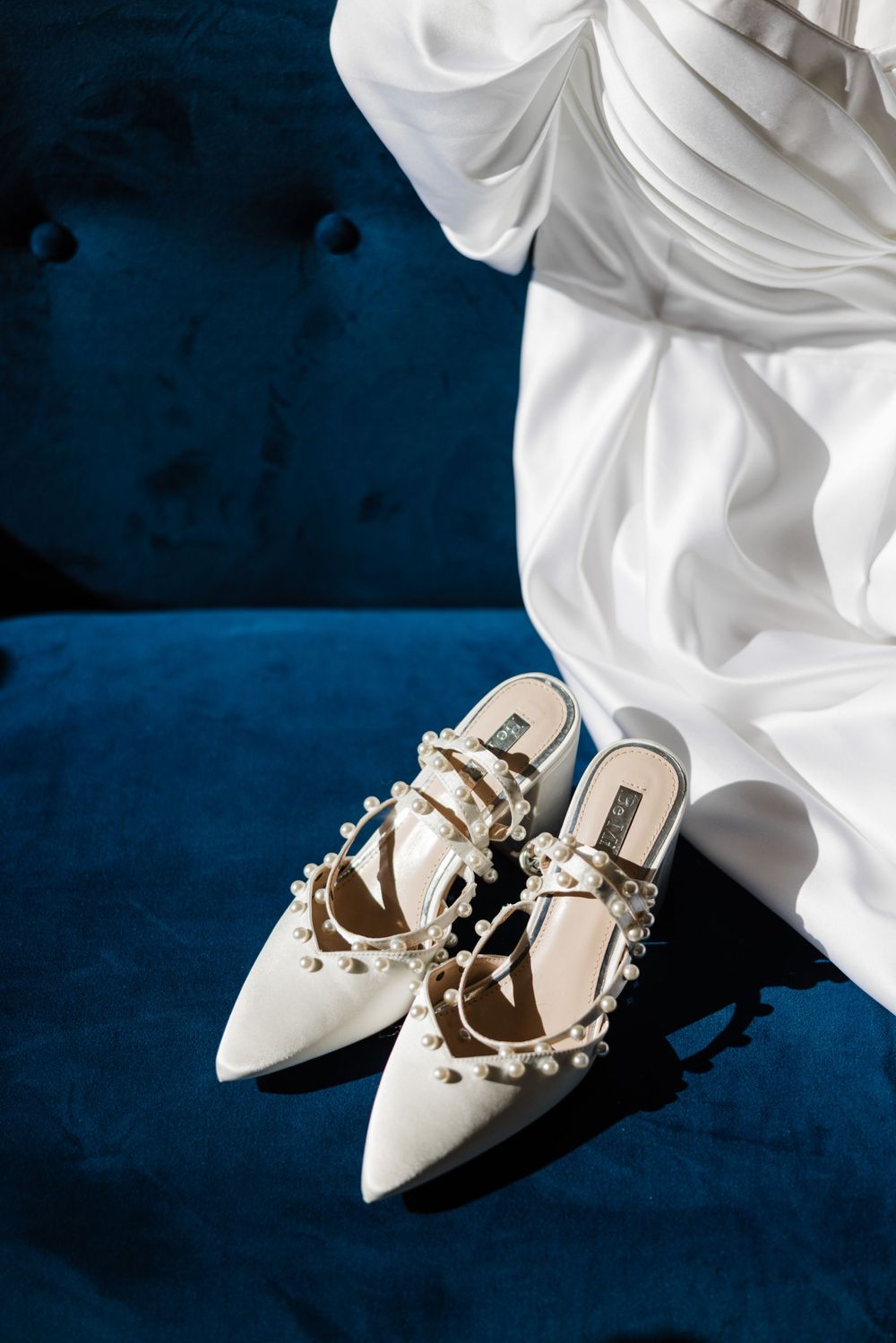 White studded bridal shoes on a navy blue velvet surface with flowing white fabric.