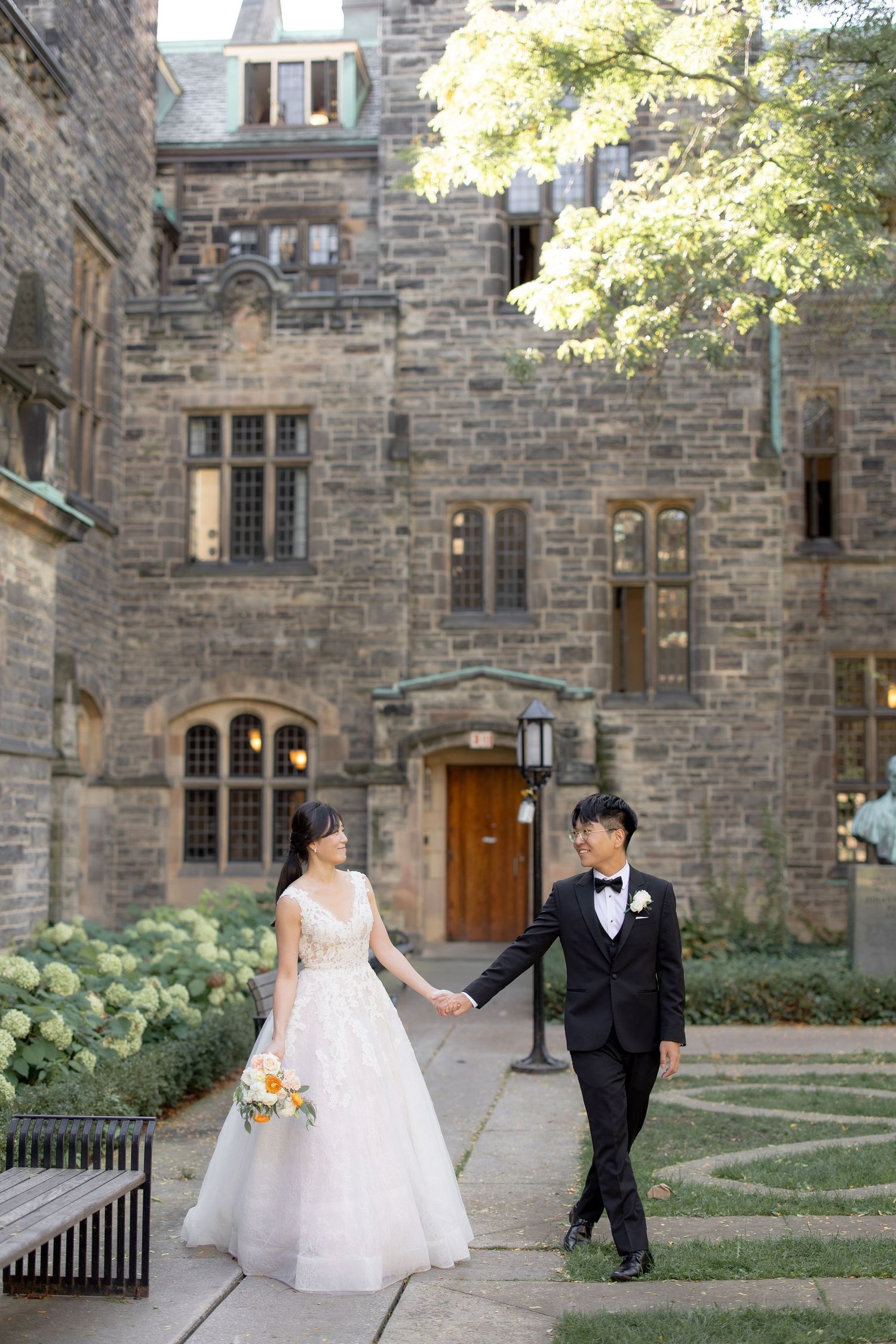 A couple walks hand in hand along a path beside a historic stone building.