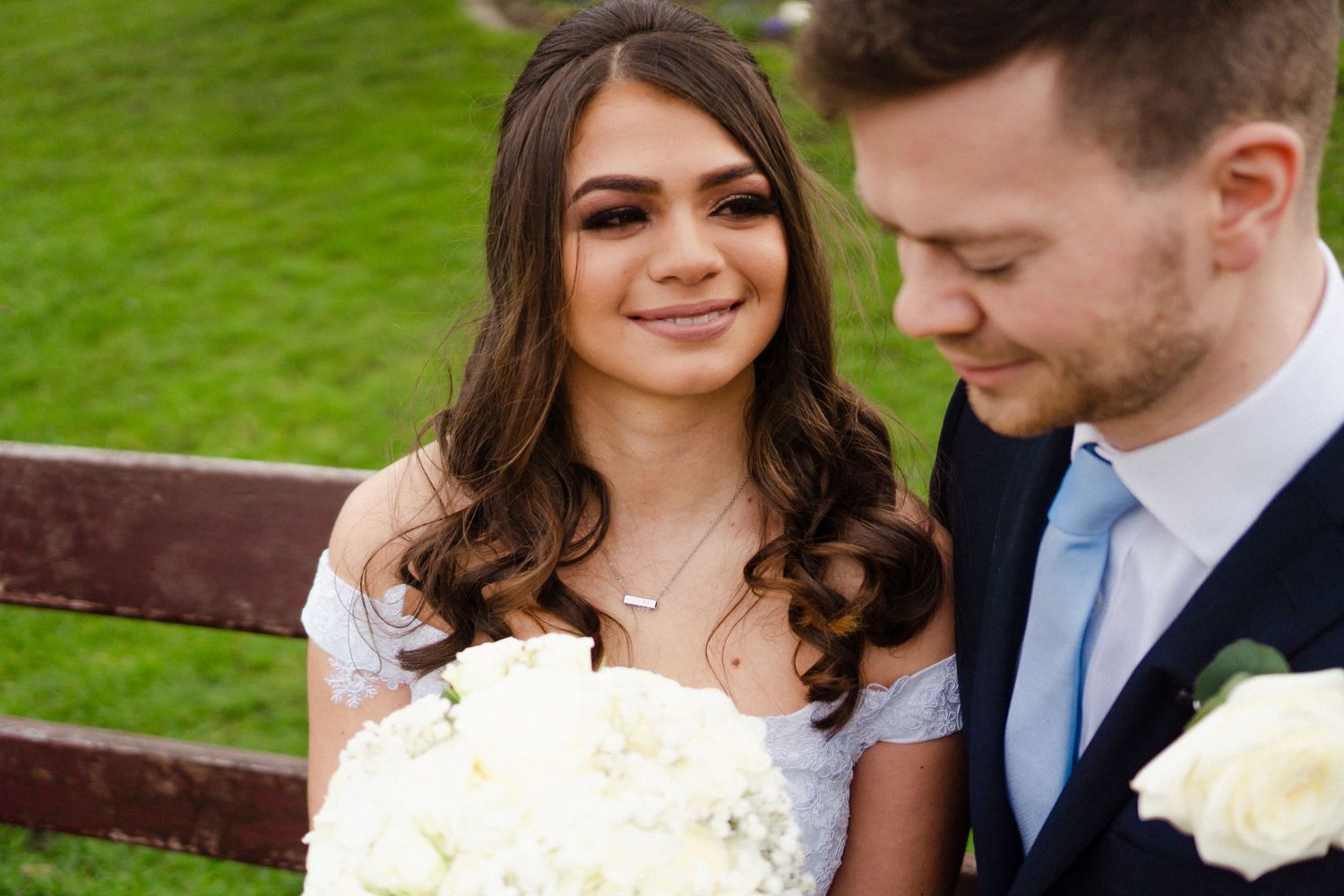 Newlyweds seated on a bench she's looking at him and he's looking down, she has a white bouquet