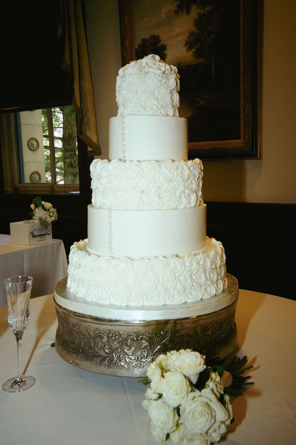 A five-tier white wedding cake with intricate lace detailing sits on an ornate silver stand next to white roses and champagne.