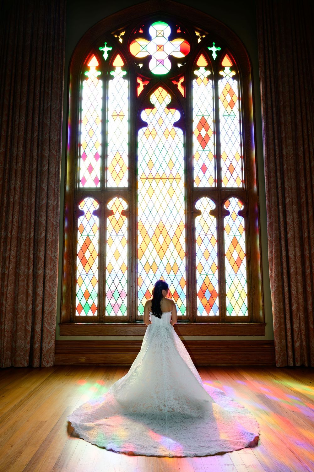 Bride in white gown with long train stands before colorful stained glass church window, light casting rainbow hues on wood floor.