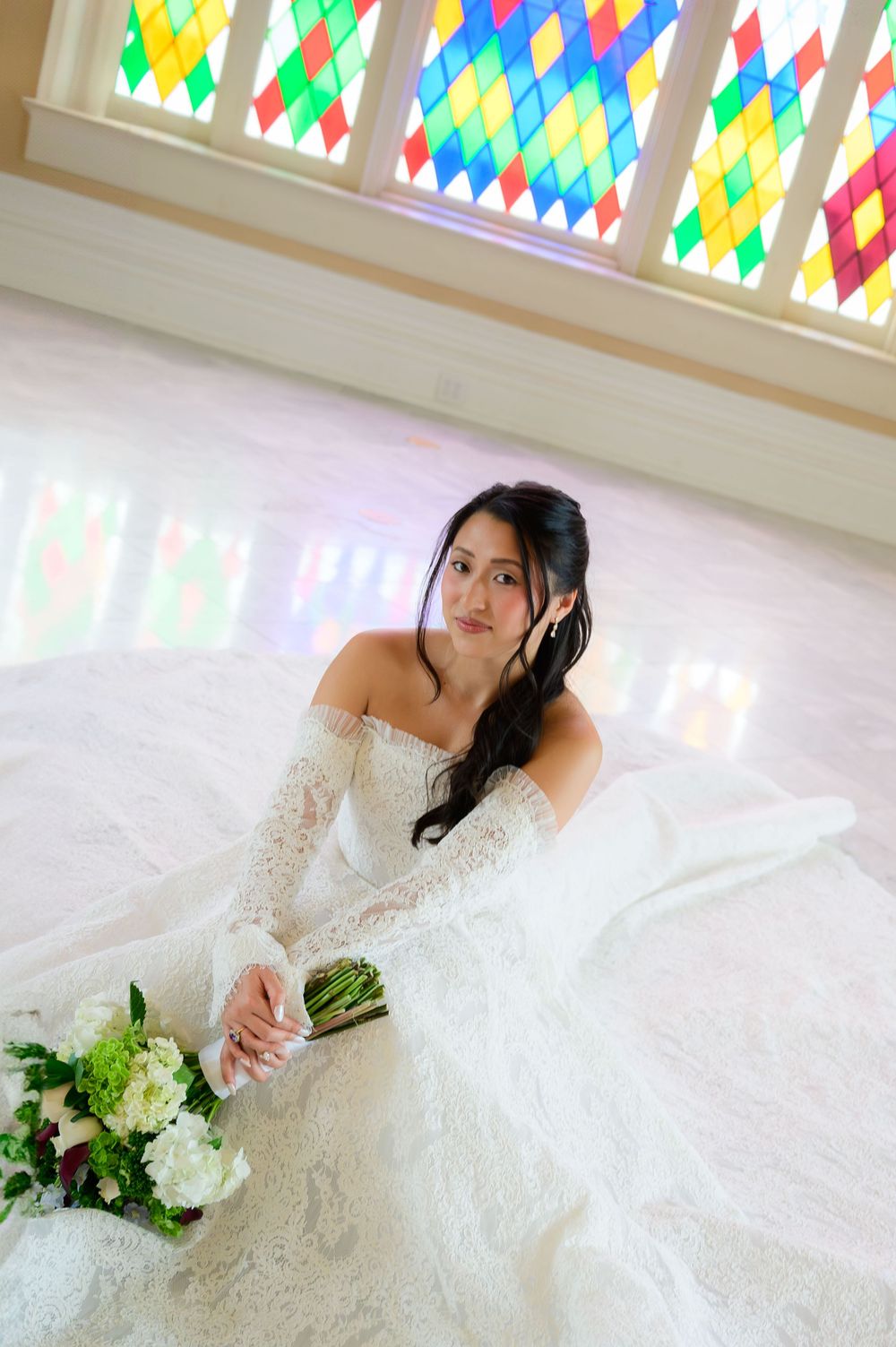 Bride in white lace off-shoulder gown sits on floor holding bouquet, colorful stained glass windows behind her.