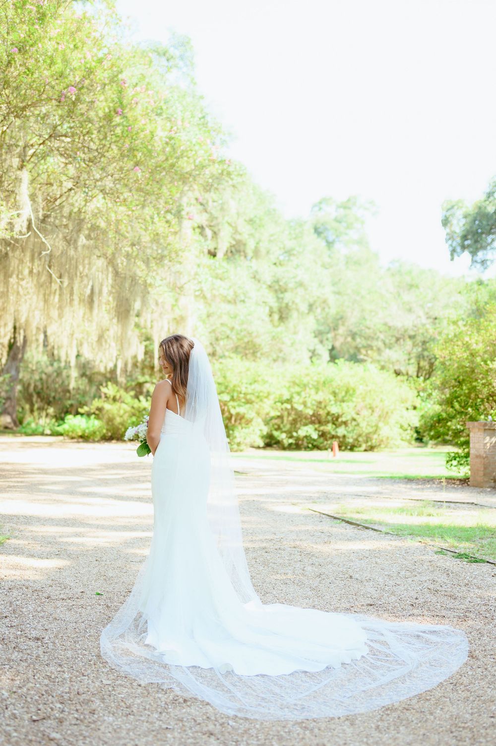 A bride in a flowing white gown with lace trim and long veil stands in a sunlit garden path surrounded by mossy trees.